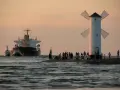 Statek i holowniki wpływają do portu obok latarni w kształcie wiatraka, na brzegu stoją ludzie.
