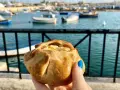 Ręka trzymająca pieczone ciastko z serem na tle maltańskiego portu z łodziami.