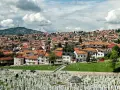 Widok na cmentarz i miasto z dachami z czerwonej dachówki pod zachmurzonym niebem.