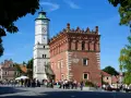 Ratusz w Sandomierzu z zegarem i zegarem słonecznym, na tle błękitnego nieba i drzew.