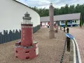 Miniatury latarni morskich na żwirowym placu, z budynkiem i łodzią w tle.