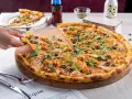 Pizza z krewetkami, oliwkami i kaparami na drewnianej desce, ręka podnosi kawałek.