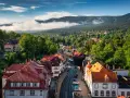 Widok z góry na miasteczko z czerwonymi dachami, otoczone zielonymi lasami i górami w tle.