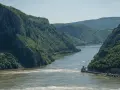 Szeroka rzeka przepływająca przez skaliste, zalesione góry. Widoczne łodzie i budynek.