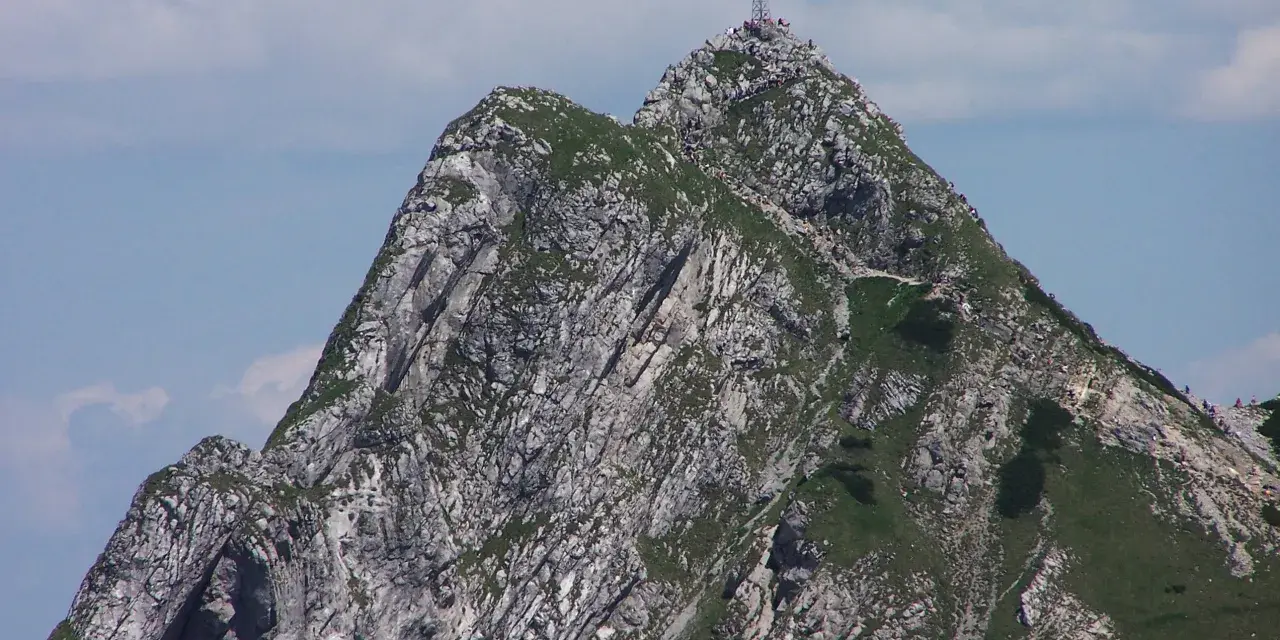Giewont z krzyżem na szczycie, skaliste zbocza, zielona trawa, błękitne niebo z chmurami.