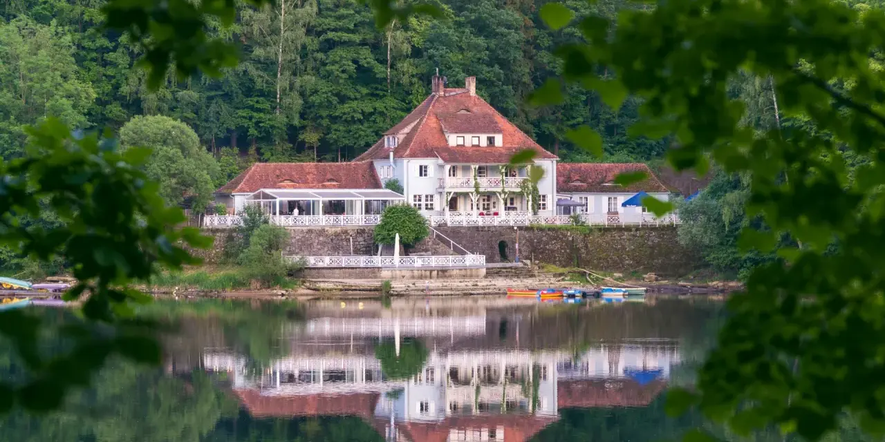 Dom z czerwonym dachem nad jeziorem, odbijający się w wodzie, otoczony zielenią.