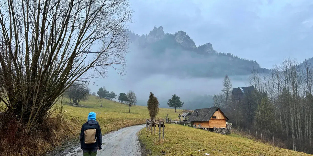 Osoba w niebieskiej czapce idzie drogą w Pieninach, w tle góry i drewniane chatki.