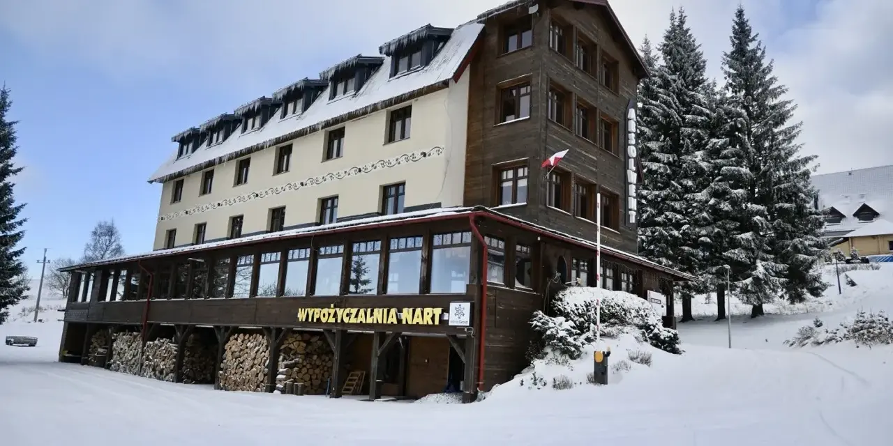 Hotel w Zieleńcu zimą, z widoczną wypożyczalnią nart i stosem drewna.