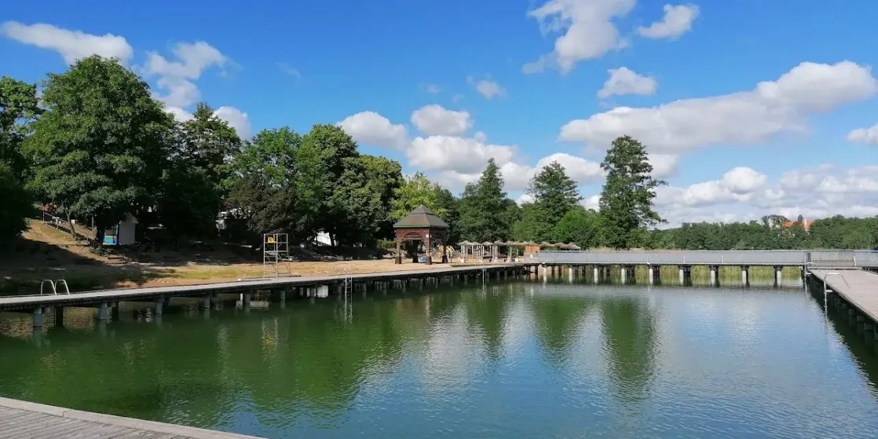 Drewniany pomost nad zieloną wodą jeziora Czos, z altaną i drzewami w tle, pod błękitnym niebem z chmurami.