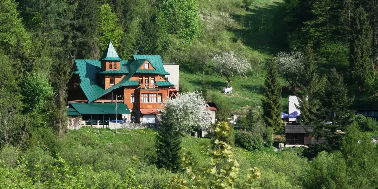 Drewniany schronisko PTTK z zielonym dachem otoczone lasem i zielonymi wzgórzami.