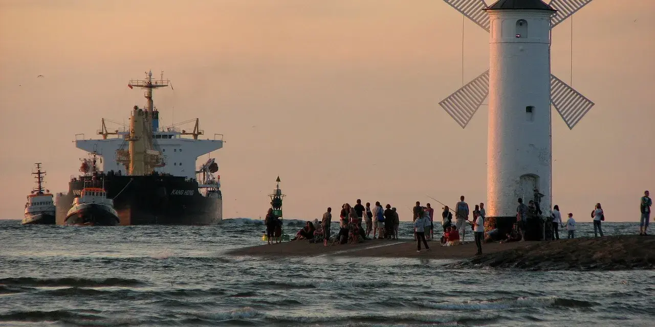 Statek i holowniki wpływają do portu obok latarni w kształcie wiatraka, na brzegu stoją ludzie.