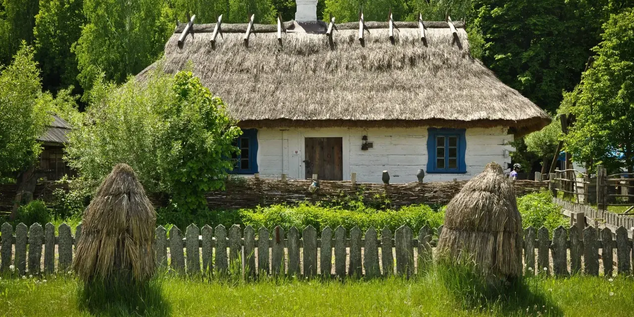 Tradycyjny biały dom z krytym strzechą dachem, otoczony drewnianym płotem i zielenią.