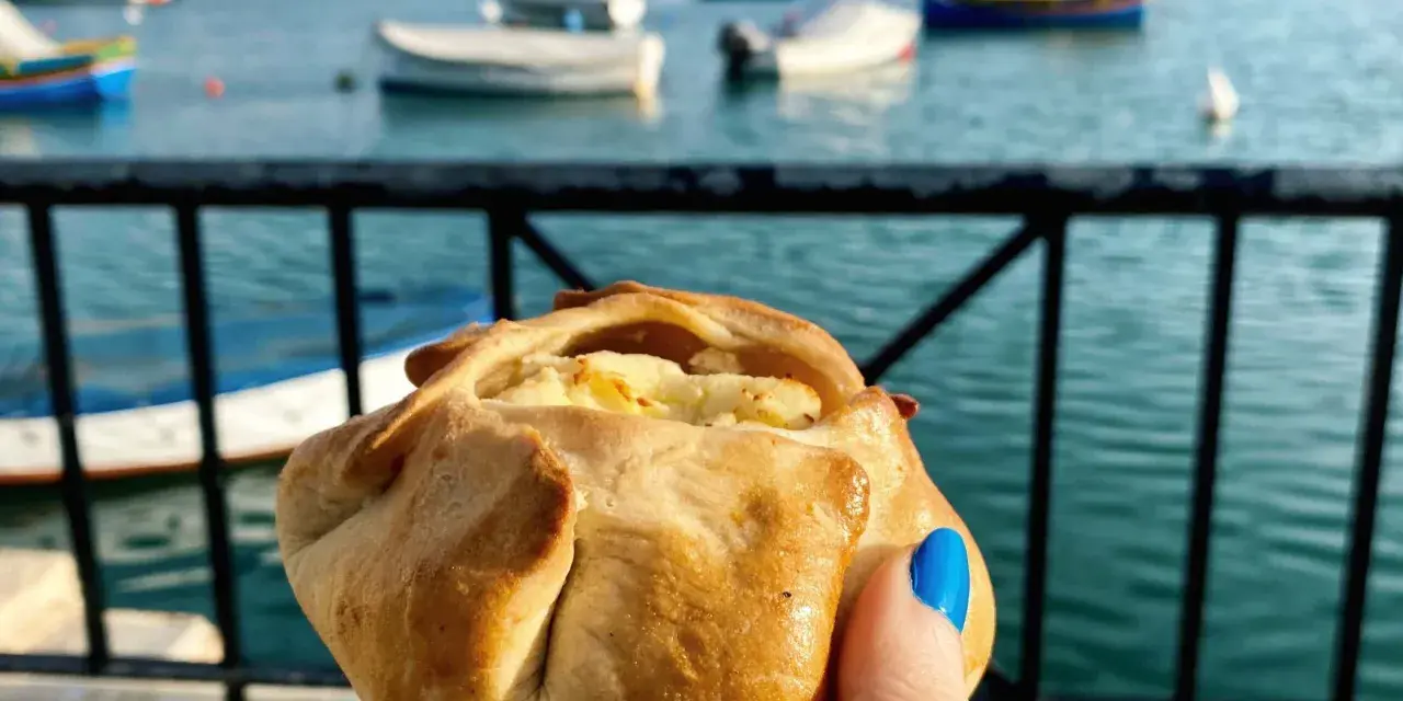 Ręka trzymająca pieczone ciastko z serem na tle maltańskiego portu z łodziami.