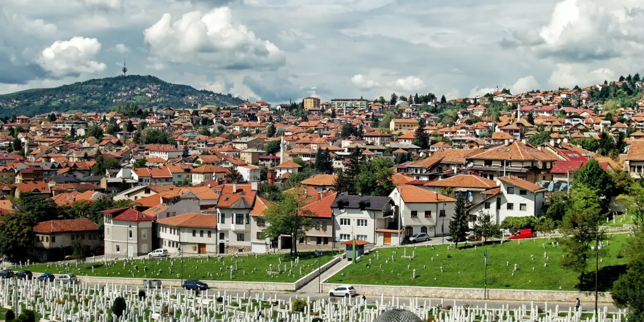 Widok na cmentarz i miasto z dachami z czerwonej dachówki pod zachmurzonym niebem.