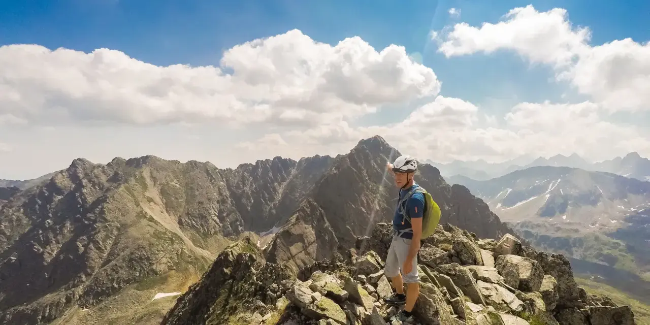 Mężczyzna w kasku i plecaku na skalistym szczycie górskim, wskazujący palcem na krajobraz.