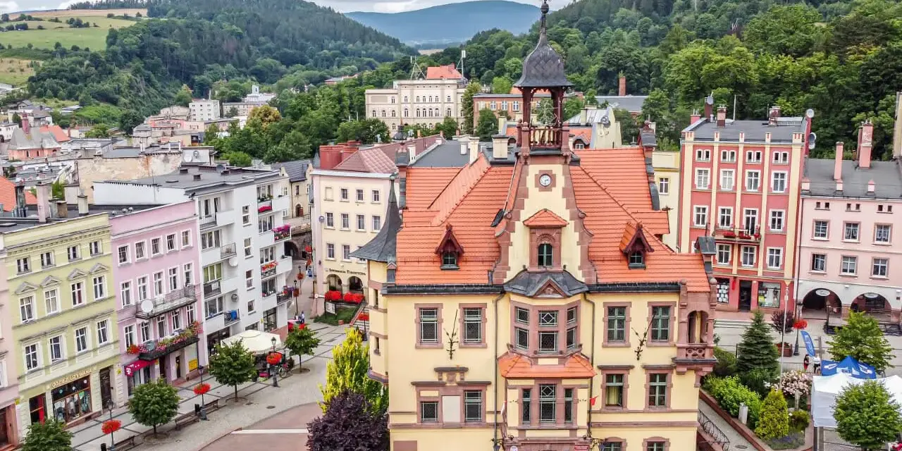 Widok na rynek w Nowej Rudzie z kolorowymi kamienicami i ratuszem w tle, otoczony wzgórzami.
