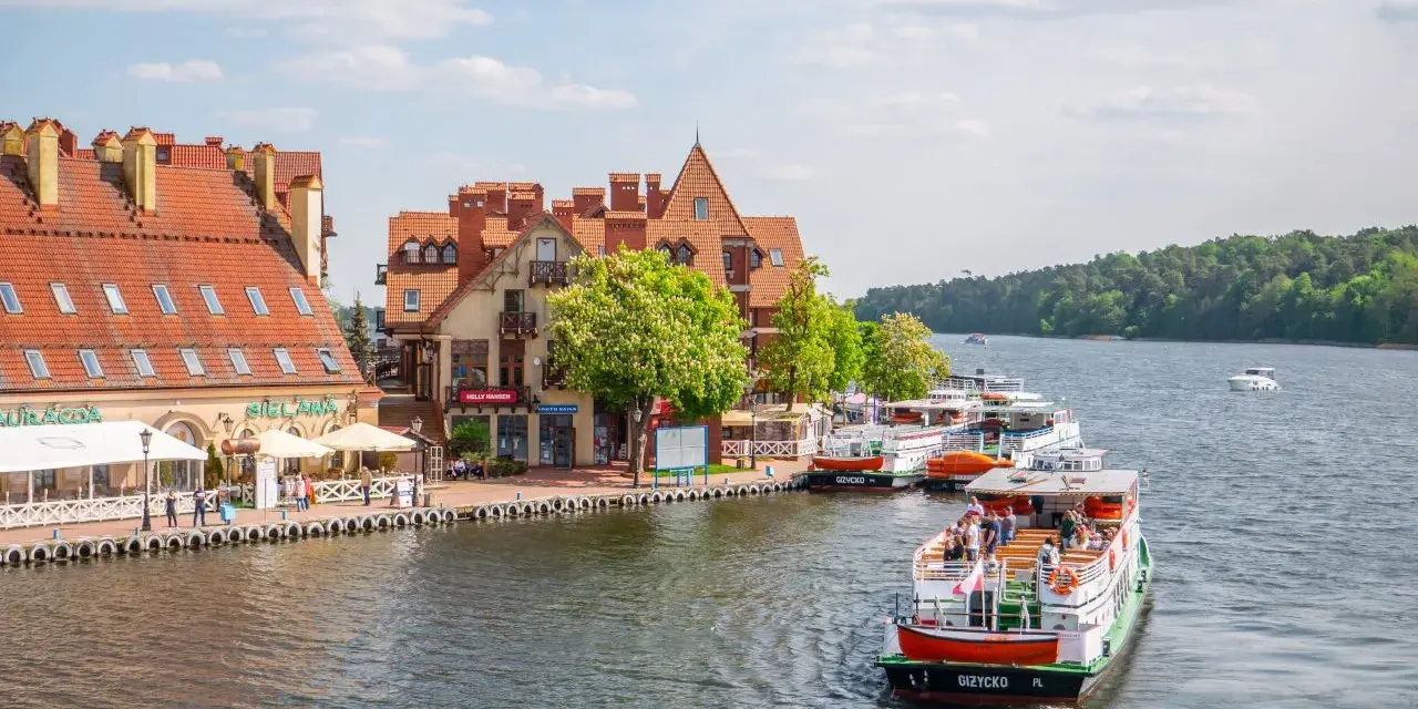 Port w Mikołajkach z zacumowanymi statkami wycieczkowymi i budynkami z czerwonymi dachami. Wspaniałe atrakcje w Mikołajkach czekają na turystów.