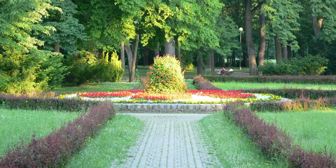 Kamienista ścieżka prowadzi przez park z kwitnącymi rabatami kwiatowymi i zielenią.