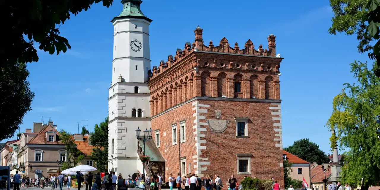 Ratusz w Sandomierzu z zegarem i zegarem słonecznym, na tle błękitnego nieba i drzew.