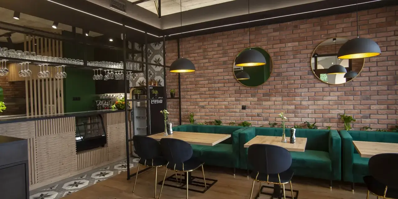 Nowoczesne wnętrze restauracji z ceglaną ścianą, zielonymi kanapami i drewnianymi stołami.