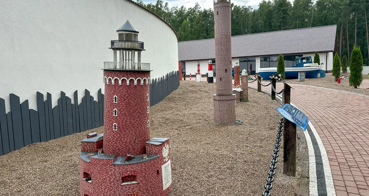 Miniatury latarni morskich na żwirowym placu, z budynkiem i łodzią w tle.