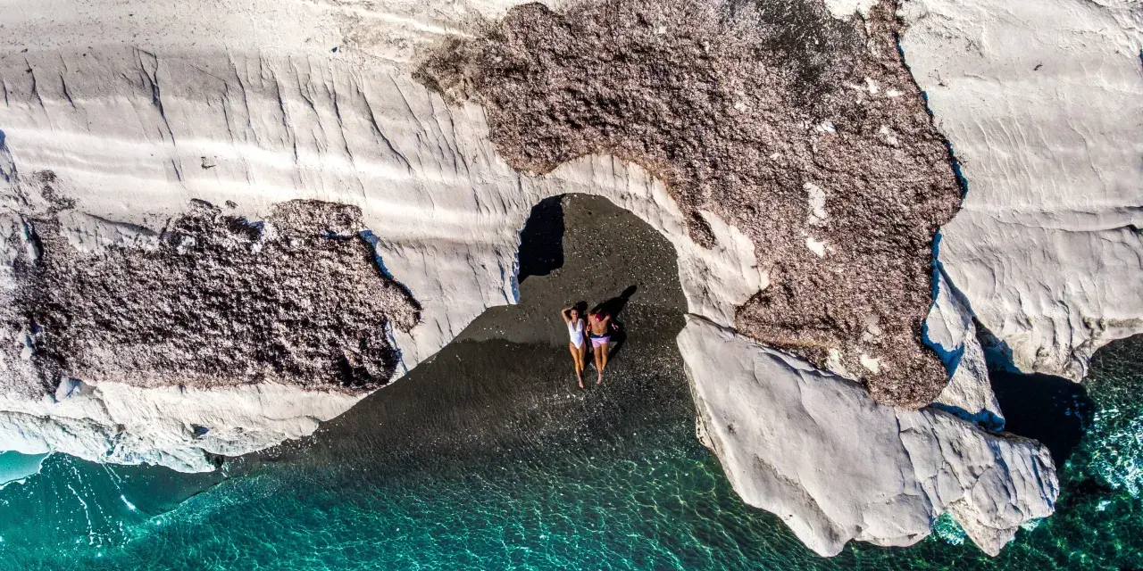 Dwie osoby leżą na czarnej plaży pod skalnym łukiem, obok turkusowej wody.
