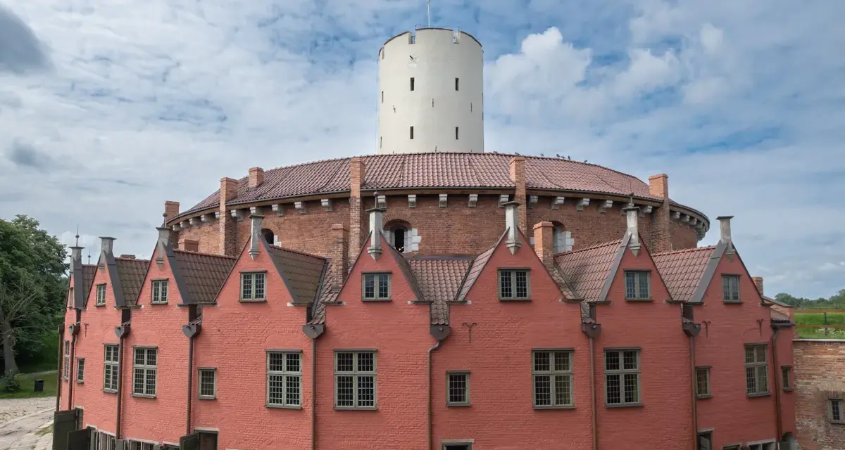 Okazały ceglany budynek z wieżą i czerwonymi domkami w kształcie podkowy.