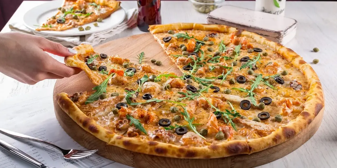 Pizza z krewetkami, oliwkami i kaparami na drewnianej desce, ręka podnosi kawałek.