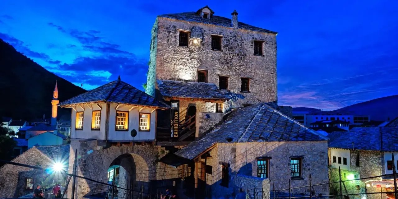 Kamienne budynki i most w Mostarze o zmierzchu, z widocznym minaretem w tle.