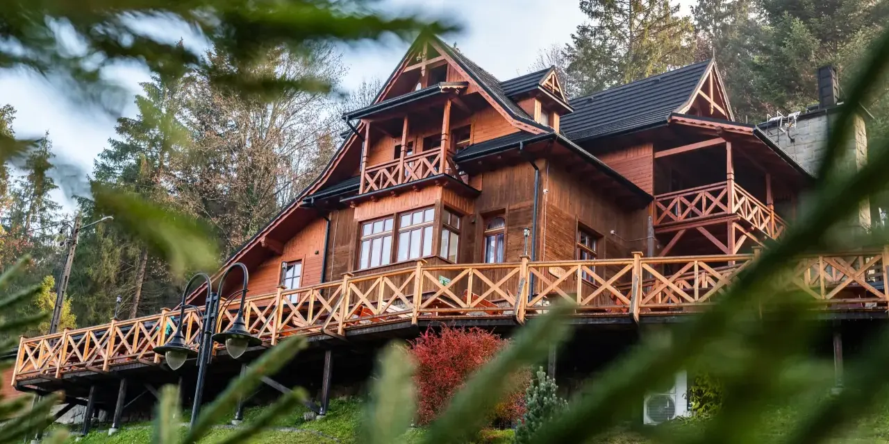 Drewniany góralski pensjonat z tarasem i balustradą, otoczony drzewami.