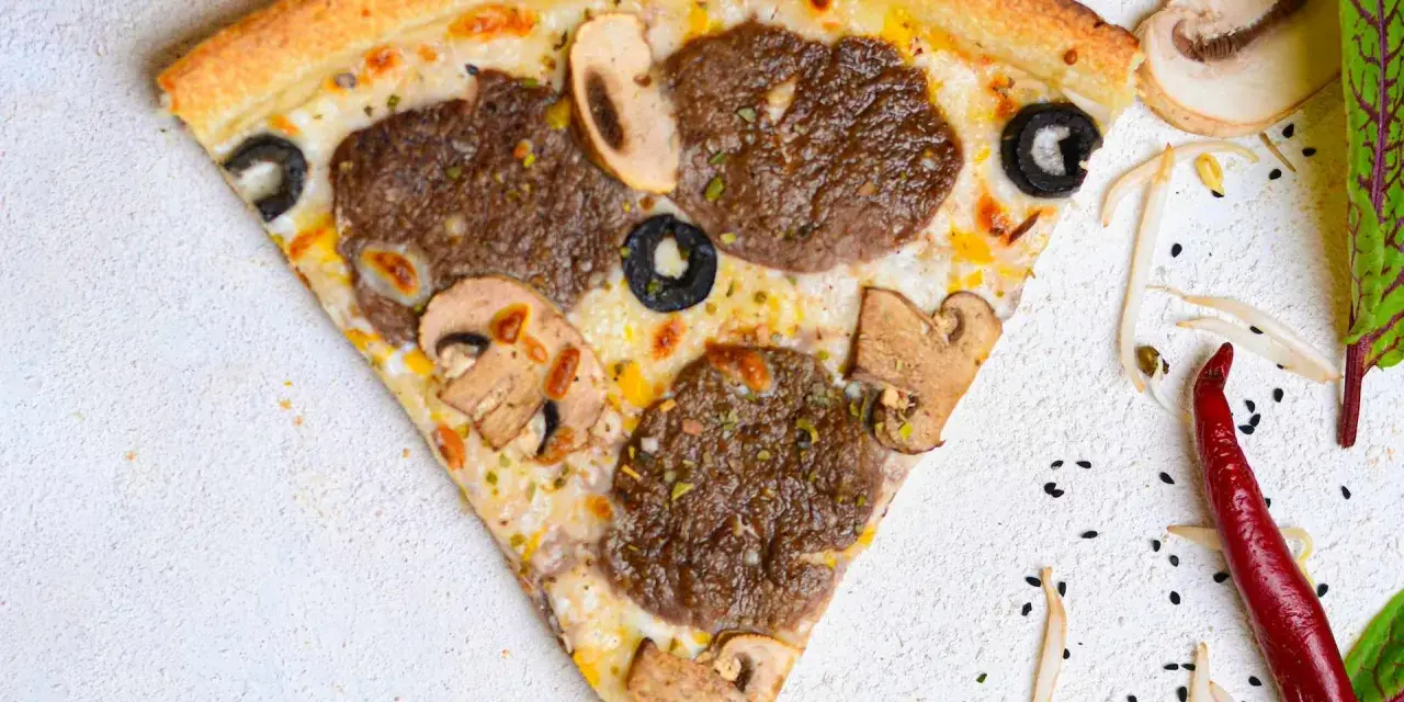 Kawałek pizzy z mięsem mielonym, pieczarkami i oliwkami na jasnym tle.