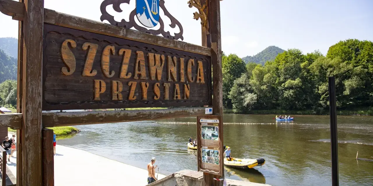 Drewniany szyld "Szczawnica Przystań" nad rzeką z tratwami i górami w tle.