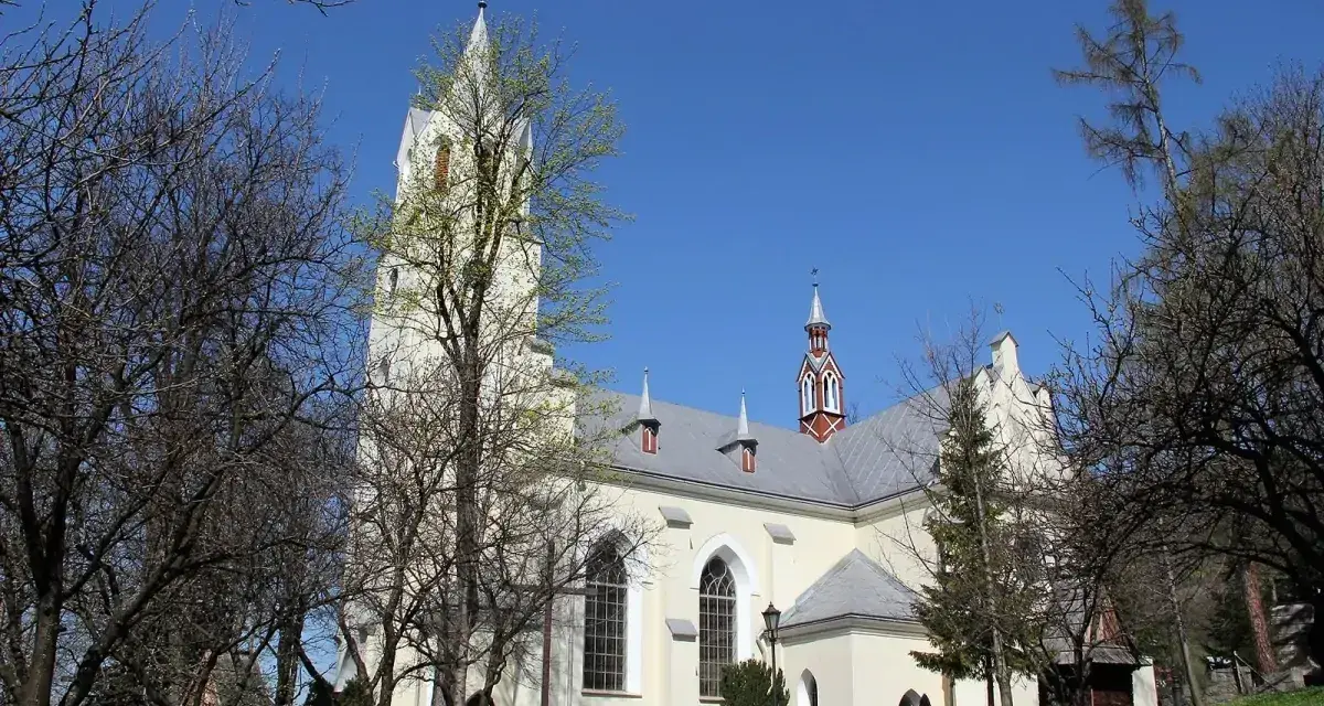 Jasny kościół św. Wojciecha w Szczawnicy z wieżą, otoczony drzewami.