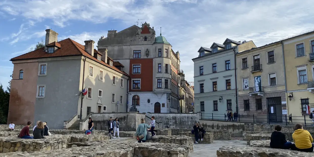 Ruiny zamku w Lublinie z ludźmi odpoczywającymi na kamiennych murach i zabytkowymi kamienicami w tle.