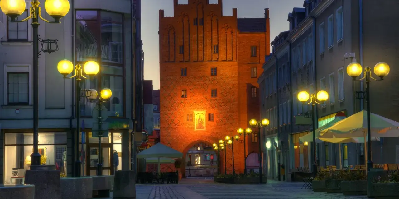 Brama miejska w Olsztynie o zmierzchu, oświetlona latarniami i iluminacją.