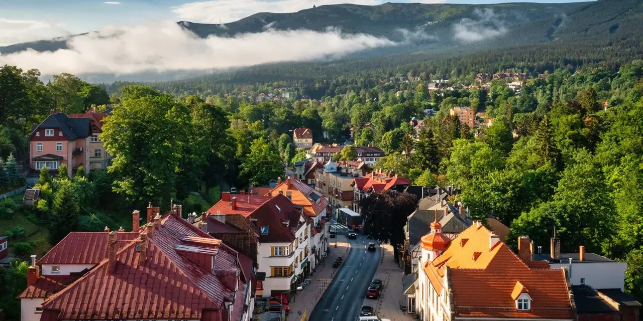 Widok z góry na miasteczko z czerwonymi dachami, otoczone zielonymi lasami i górami w tle.
