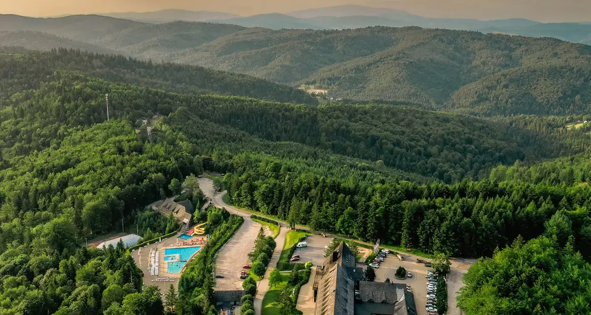 Widok z lotu ptaka na hotel SPA z basenem i zjeżdżalnią, otoczony lasem i górami.