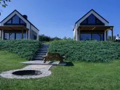 Dwa nowoczesne domki z dużymi oknami na zielonym wzgórzu, z miejscem na ognisko i stołem piknikowym.
