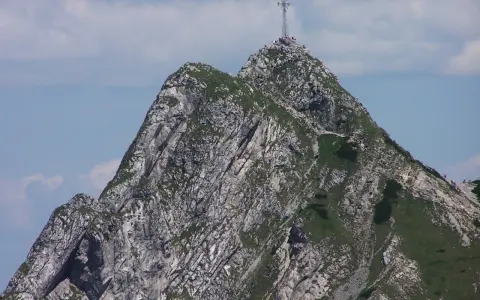 Giewont z krzyżem na szczycie, skaliste zbocza, zielona trawa, błękitne niebo z chmurami.