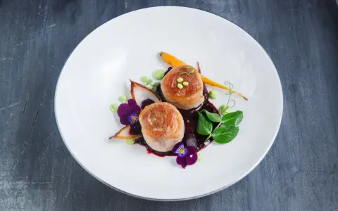 Eleganckie danie z dwóch medalionów mięsa zawiniętych w boczek, podane z purée, marchewką i kwiatami.