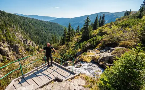 Mężczyzna na drewnianej platformie z widokiem na górski krajobraz i wodospad.