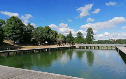 Drewniany pomost nad zieloną wodą jeziora Czos, z altaną i drzewami w tle, pod błękitnym niebem z chmurami.
