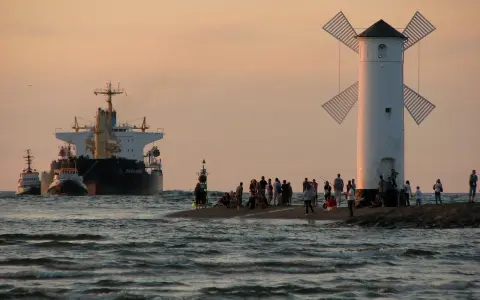 Statek i holowniki wpływają do portu obok latarni w kształcie wiatraka, na brzegu stoją ludzie.