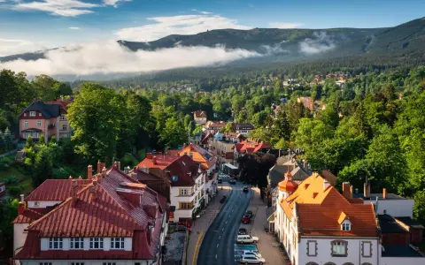 Widok z góry na miasteczko z czerwonymi dachami, otoczone zielonymi lasami i górami w tle.
