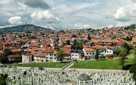 Widok na cmentarz i miasto z dachami z czerwonej dachówki pod zachmurzonym niebem.