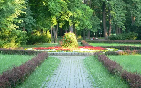 Kamienista ścieżka prowadzi przez park z kwitnącymi rabatami kwiatowymi i zielenią.