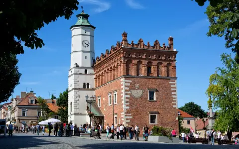 Ratusz w Sandomierzu z zegarem i zegarem słonecznym, na tle błękitnego nieba i drzew.