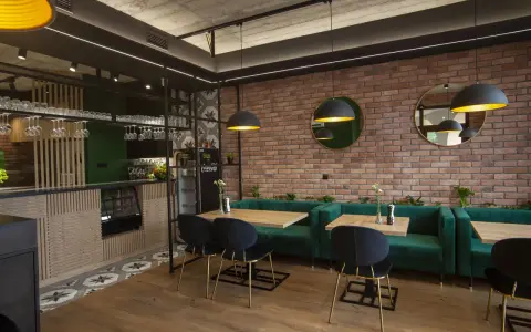 Nowoczesne wnętrze restauracji z ceglaną ścianą, zielonymi kanapami i drewnianymi stołami.
