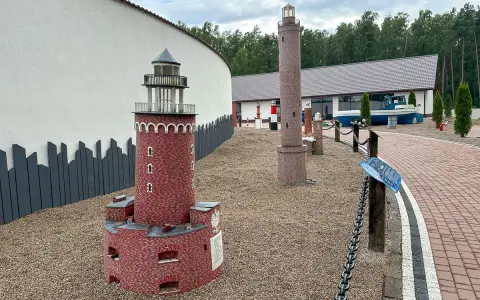 Miniatury latarni morskich na żwirowym placu, z budynkiem i łodzią w tle.