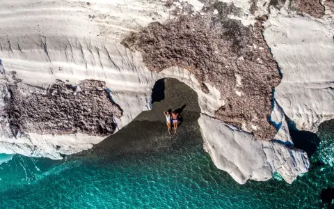 Dwie osoby leżą na czarnej plaży pod skalnym łukiem, obok turkusowej wody.